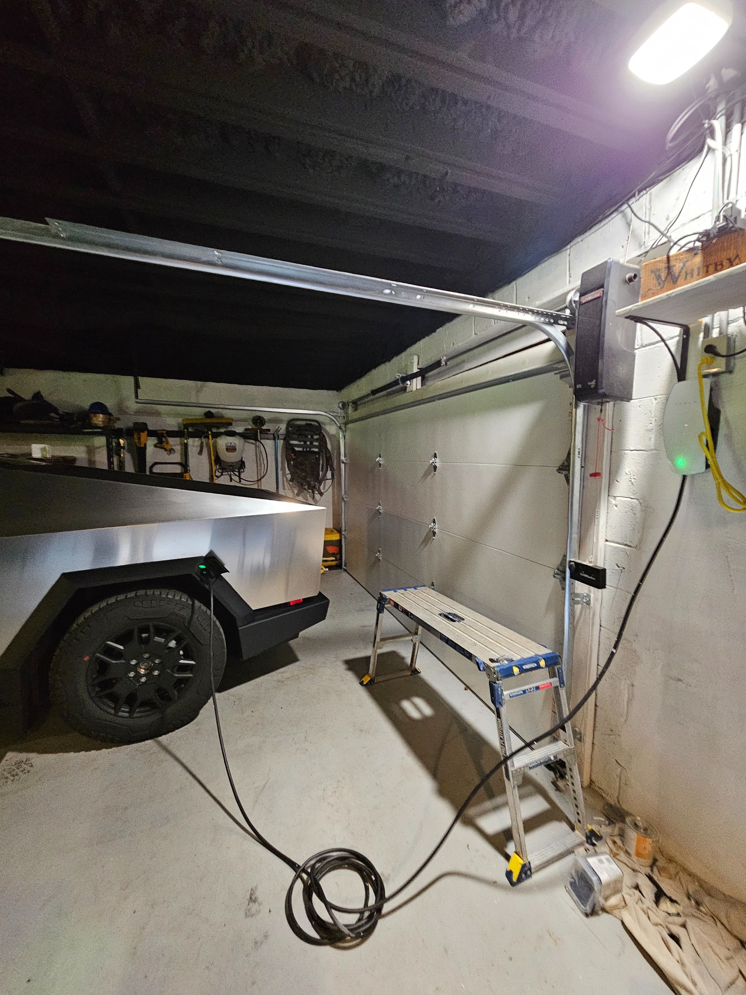 A futuristic vehicle parked in a garage, with a step ladder nearby and a coiled cable hanging from the wall.