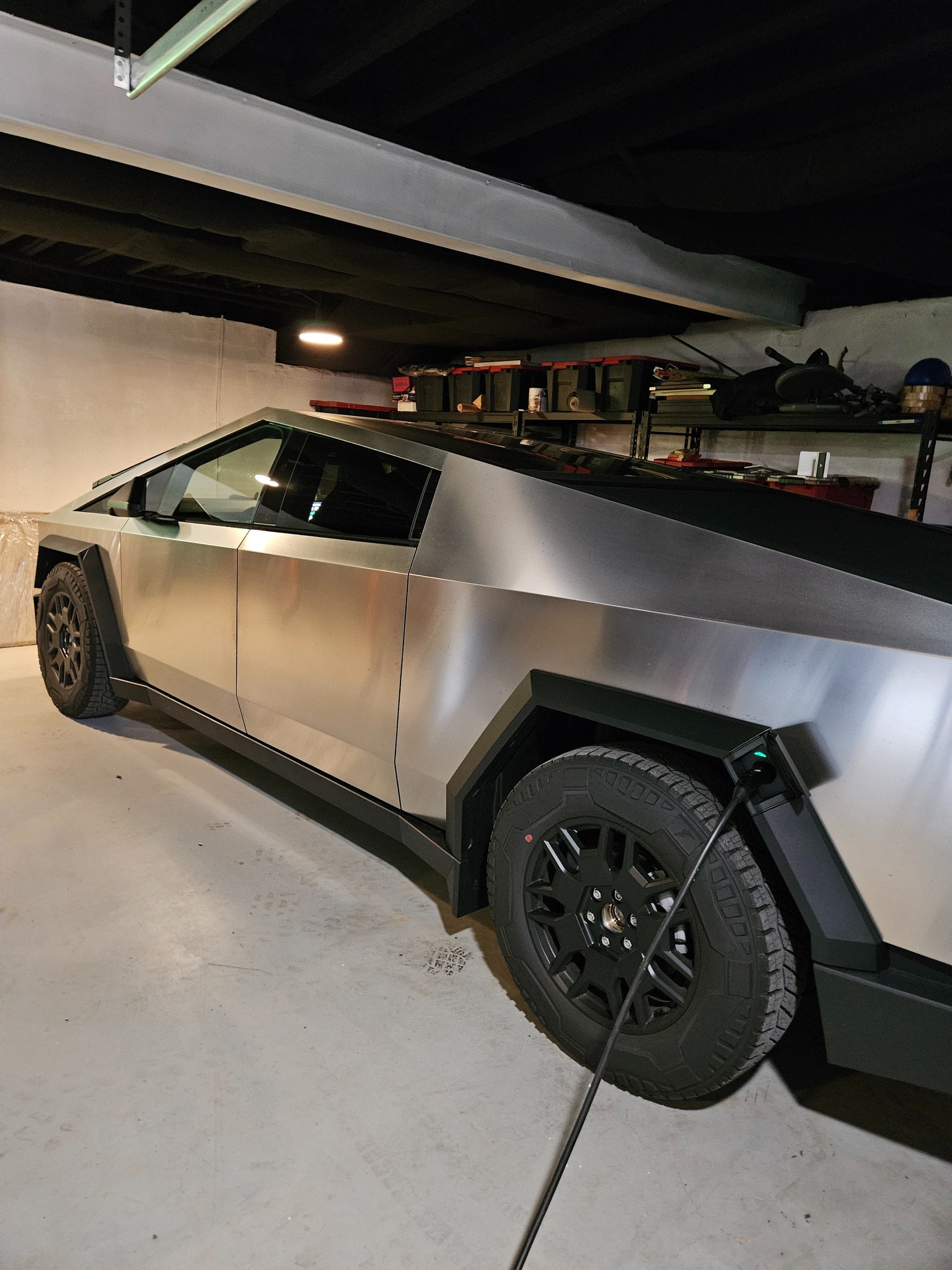 A futuristic, angular metallic vehicle parked in a garage, viewed from the side, with a cable connected to it. Shelving and storage items are visible in the background.