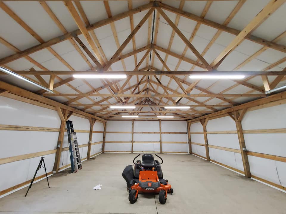 Spacious wooden garage interior with a lawn mower centered on the concrete floor. A ladder and tripod are positioned against the wall. White ceiling panels are supported by wooden beams.