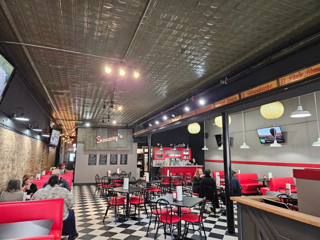 A retro-style diner with red booths, black and white checkered flooring, and several patrons seated. A TV is mounted on the wall, and the ceiling has exposed bulbs.