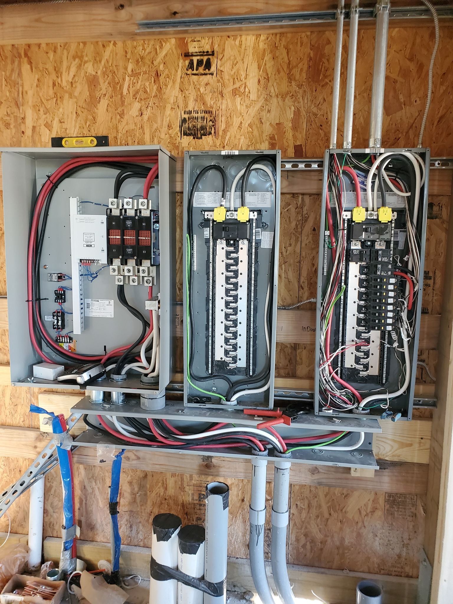 Three electrical breaker panels with various colored wires and switches mounted on a wooden wall. Pipes are visible below the panels.