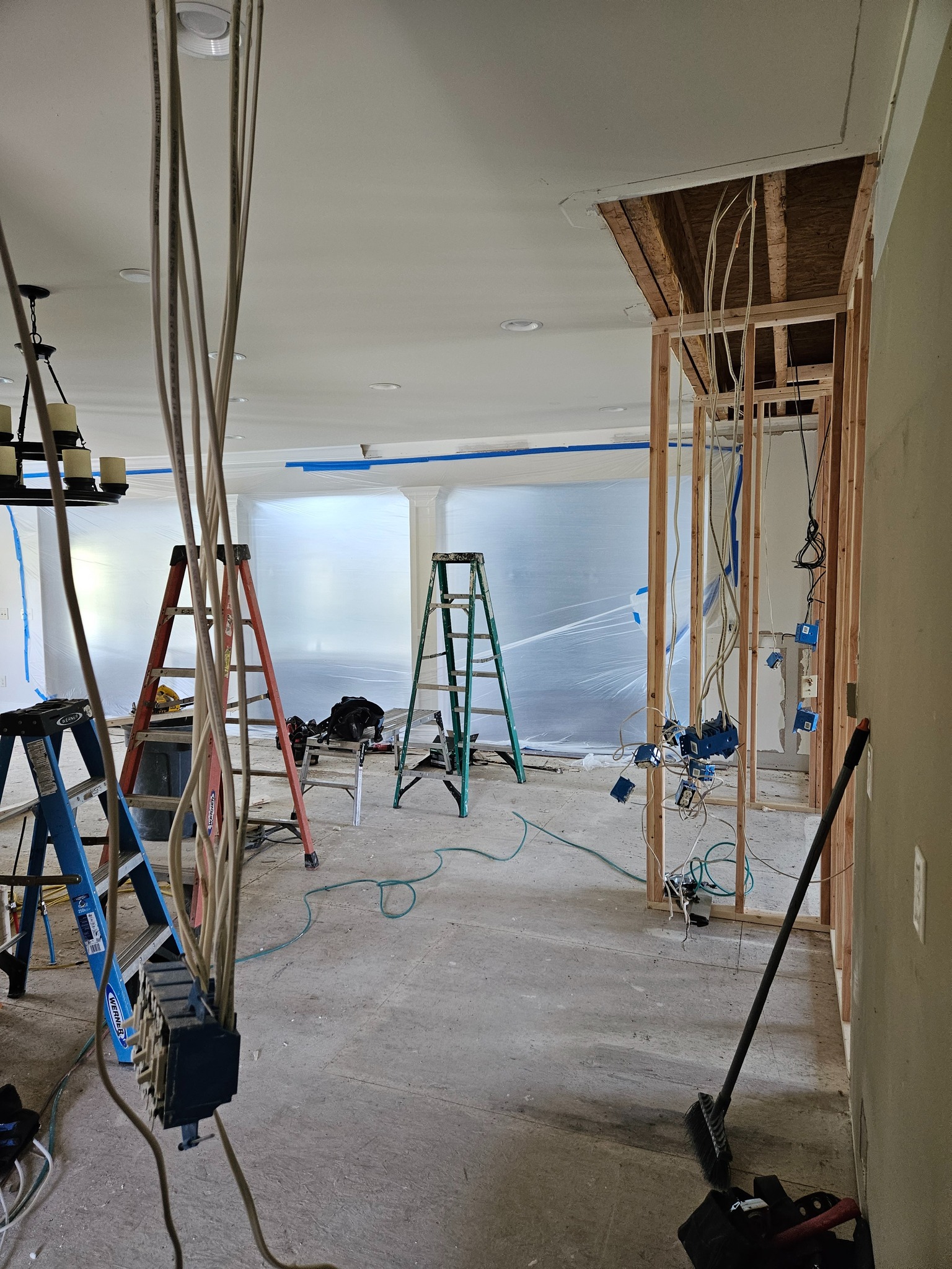 Room under renovation with exposed wires, ladders, and tools. Ceiling partially removed and plastic sheeting covers windows.