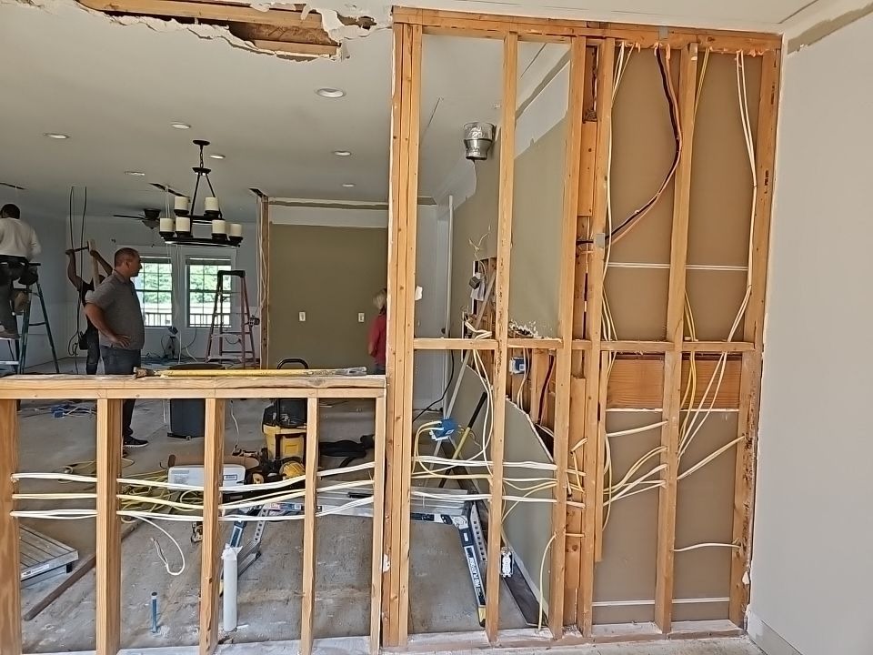 Interior renovation scene with exposed wooden framework and electrical wiring. Workers install ceiling lights in a partially finished room.